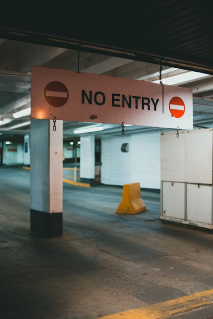 A white rectangular 'NO ENTRY' sign with black text and red circular icons on each side is suspended from the ceiling of an underground car park or loading area. The sign hangs above a concrete pillar and a small yellow wheel stop positioned on a dark grey asphalt surface. In the background, there are white-painted walls, structural supports, and a partially visible vehicle lane, indicating the space is used for vehicle access and loading activities. The environment is lit with artificial lighting, emphasizing the area as a potential site for house removals or furniture transport, with Knightsbridge Movers providing professional moving services such as packing, loading, and home relocation logistics within this setting.