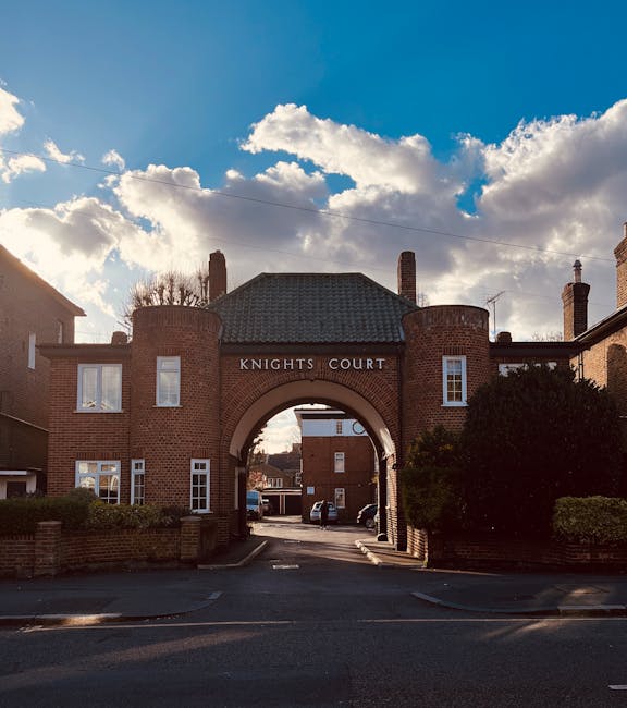 View of Knights Court, a brick residential building with a central archway entrance, situated inside a property boundary with surrounding trees and bushes. The archway opens onto a driveway where several cardboard moving boxes and a large wrapped piece of furniture are being carried or moved by workers engaged in home relocation activities. Visible packaging materials include plastic wrap and blankets used to protect furniture during furniture transport and packing. A moving trolley or equipment may be nearby, supporting the loading process. The background shows a partly cloudy sky with sunlight casting shadows on the premises, and the scene suggests an ongoing house removal operation coordinated by Knightsbridge Movers, specializing in removals and packing services for house relocations within Knightsbridge.