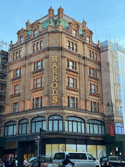 A historic multi-storey building with ornate architectural details and a decorative green rooftop. The facade features large arched windows on the ground floor, with the name 'HARRUDS' displayed vertically in gold lettering on the corner of the building. In the foreground, there are parked cars and a white van along a busy street, with pedestrians walking on the pavement. The image depicts a busy urban environment typical of central London, relevant to house removals and moving services such as those offered by Knightsbridge Movers, especially for flats on Brompton Road, highlighting the typical setting for furniture transport and home relocation activities.