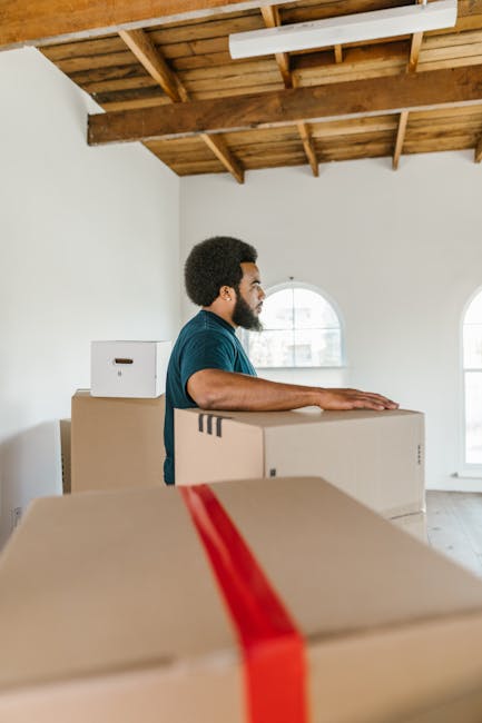 A man with dark curly hair and a beard, dressed in a blue T-shirt, is seen in a bright, spacious room with high wooden-beamed ceilings and large arched windows that let in natural light. He is standing behind several packed cardboard boxes, which are sealed with red and brown packing tape, and appears to be preparing for a home relocation or furniture transport. The boxes vary in size and are positioned on the floor, with some stacked. The environment has a minimalistic, clean aesthetic with white walls and minimal visible furniture or decor. The open space and visible window frames suggest the interior of a flat or apartment being prepared for moving, and the scene illustrates the packing and loading process involved in professional removals, like those offered by Knightsbridge Movers, supporting a seamless relocation service.