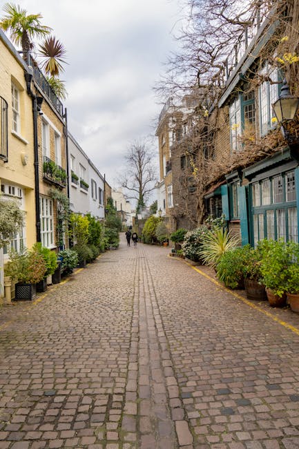 A cobbled street in a residential area with mews houses on either side, featuring a mix of traditional brick and modern painted facades. Potted plants and greenery border the footpath, with some trees visible in the distance under an overcast sky. In the foreground, a group of movers from Knightsbridge Movers are engaged in a home relocation process, loading cardboard boxes and wrapped furniture onto a van parked on the street. The items are carefully packed with plastic wrap and blankets, and the movers are using trolleys and straps to facilitate furniture transport. The scene highlights the interior of a property near the street, where furniture and boxes are ready to be loaded for a house removals service, illustrating the physical process of packing and loading during a residential move in London.