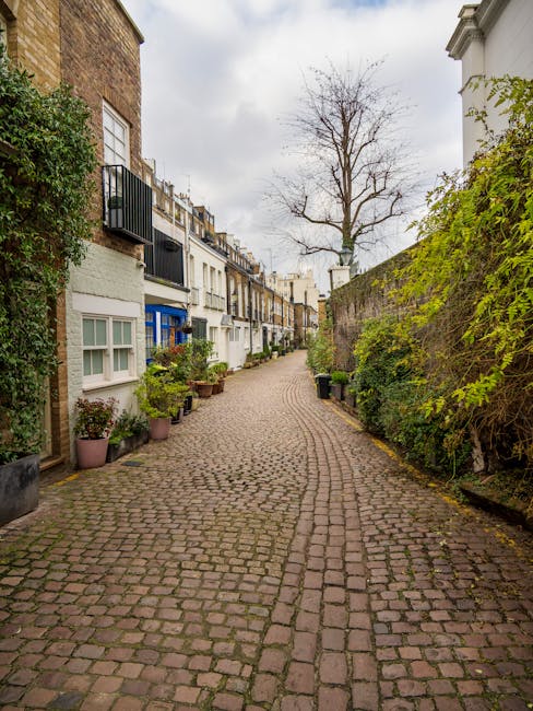 A cobbled street in a residential area with mews houses on either side, featuring a mix of traditional brick and modern painted facades. Potted plants and greenery border the footpath, with some trees visible in the distance under an overcast sky. In the foreground, a group of movers from Knightsbridge Movers are engaged in a home relocation process, loading cardboard boxes and wrapped furniture onto a van parked on the street. The items are carefully packed with plastic wrap and blankets, and the movers are using trolleys and straps to facilitate furniture transport. The scene highlights the interior of a property near the street, where furniture and boxes are ready to be loaded for a house removals service, illustrating the physical process of packing and loading during a residential move in London.