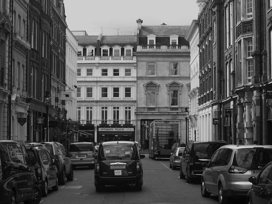 Black and white photograph of a narrow urban street in Knightsbridge with tightly parked cars lining both sides of the road. In the foreground, a small black van is seen, likely used for home relocation and furniture transport, positioned close to the curb on the left. Behind it, various vehicles including hatchbacks, sedans, and a larger van are parked along the street, which is adjacent to ornate, historic-style buildings with large windows, decorative mouldings, and mansard roofs. Some of these vehicles are partially visible, indicating limited space for loading and unloading activities typical of house removals in busy city areas. Visible in the distance is a delivery truck or moving van already partially loaded or parked near a building entrance, suggesting ongoing or upcoming packing and moving operations. The environment appears quiet with no pedestrians, and the overall scene highlights logistical considerations for loading bays or street-based removals in a central London location, aligning with services provided by Knightsbridge Movers.