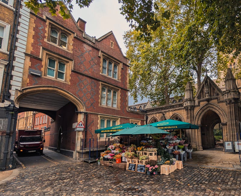 A view of a historic brick building with arched windows and an arched doorframe, characteristic of traditional architecture in the Knightsbridge area. In the foreground, a flower stall with a green canopy displays an array of colorful flowers and plants arranged in wooden crates and containers on cobblestone pavement. A small plastic container with chalkboard signs is positioned near the flowers, providing pricing or product details. To the left of the flower stall, a black van is partially visible, parked near a narrow road that passes through an arched passage underneath the building. The surrounding environment includes tall trees with green foliage, some of which extend over the building, creating a shaded area. The scene seems set during daylight hours, with natural light illuminating the textured brickwork, and the overall context suggests an urban setting conducive to home relocation or moving services, as coordinated by Knightsbridge Movers, supporting loading and transportation processes in a busy, historic part of Knightsbridge.