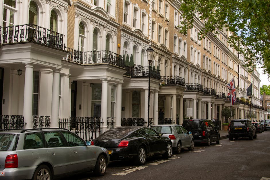 Black and white photograph of a narrow urban street in Knightsbridge with tightly parked cars lining both sides of the road. In the foreground, a small black van is seen, likely used for home relocation and furniture transport, positioned close to the curb on the left. Behind it, various vehicles including hatchbacks, sedans, and a larger van are parked along the street, which is adjacent to ornate, historic-style buildings with large windows, decorative mouldings, and mansard roofs. Some of these vehicles are partially visible, indicating limited space for loading and unloading activities typical of house removals in busy city areas. Visible in the distance is a delivery truck or moving van already partially loaded or parked near a building entrance, suggesting ongoing or upcoming packing and moving operations. The environment appears quiet with no pedestrians, and the overall scene highlights logistical considerations for loading bays or street-based removals in a central London location, aligning with services provided by Knightsbridge Movers.