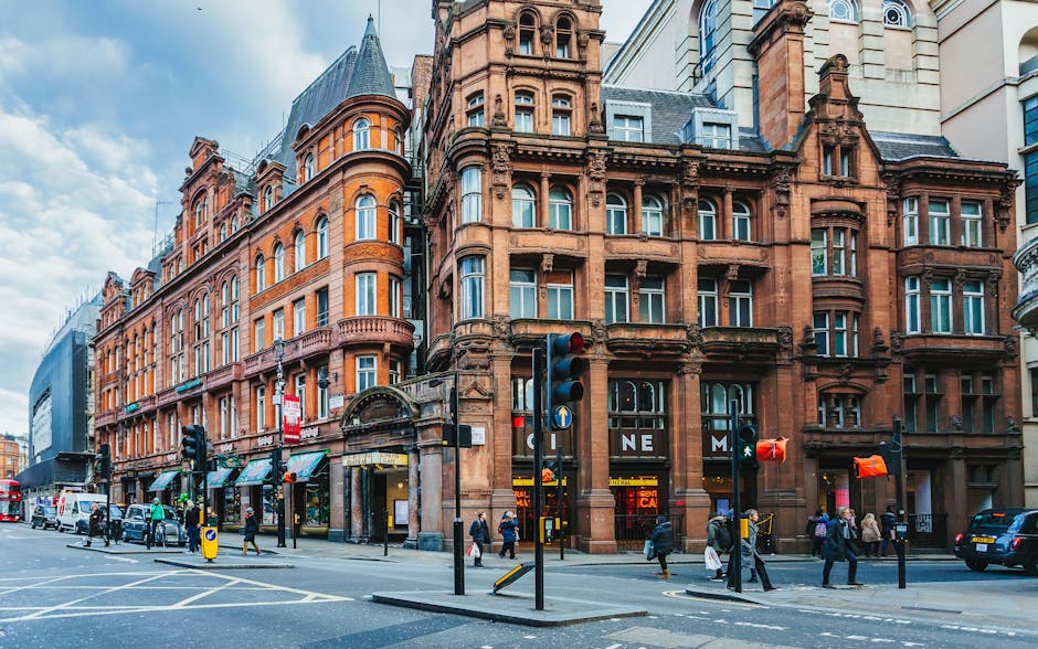 Photograph of a historic red-brick building situated on a busy street corner with multiple storefronts at the ground level. The building features ornate architectural details, rounded bay windows, and decorative columns, typical of city centre architecture. Pedestrians are walking along the sidewalk, some carrying shopping bags, while others wait at pedestrian crossings near traffic lights. Several vehicles, including cars and a delivery van, are parked or moving along the street. In the foreground, there are traffic signals, a pedestrian crossing, and a yellow bollard indicating a designated zone for loading or deliveries. The scene is well-lit with natural daylight, emphasizing the textures of the brickwork and the urban environment. This setting illustrates a typical cityscape where local removals or house relocation services by Knightsbridge Movers could operate, involving the loading and transport of household furniture and belongings between buildings in a busy residential and commercial area.