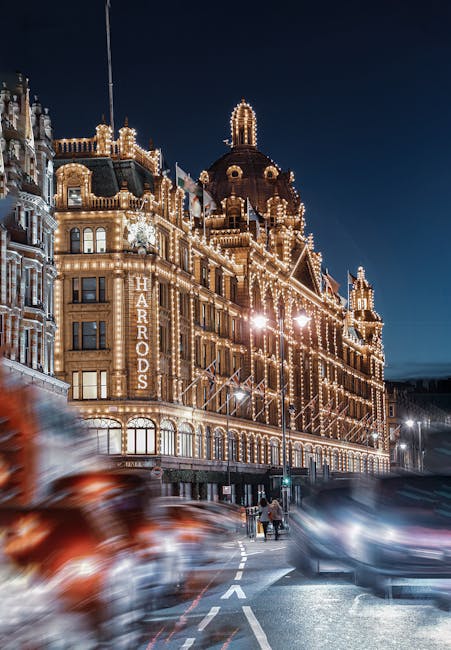A nighttime view of the historic Harrods department store in Knightsbridge, illuminated with warm golden lighting that highlights its ornate architectural details and domed roof. The building's classic façade features multiple windows and decorative elements, with the Harrods sign prominently displayed along the side. In the foreground, blurred cars move along the street, creating light streaks from their headlights, while a few pedestrians walk on the pavement near the curb. The scene captures the bustling atmosphere typical during evening hours, with the store's lighting contrasting against the dark sky. This image emphasizes the central location within Knightsbridge, suitable for illustrating home relocation or property transfer services, and the surrounding urban environment during a busy city evening. Knightsbridge Movers, specialists in removals, often operate in such iconic areas, facilitating furniture transport and packing during house moves in the vicinity.