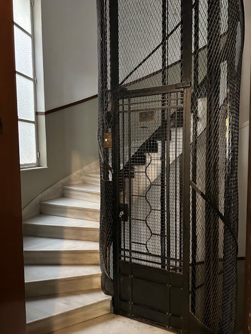 A black metal lift with a wire mesh design is positioned at the base of a staircase inside a property, serving as an access aid for home relocation or furniture transport. The lift is partially extended with its gate open, revealing a space for goods or boxes to be moved between floors. To the left of the lift, a stone staircase with polished, light-colored steps curves upward, illuminated by natural light coming through a frosted window. The interior walls are painted in a neutral tone, and a wooden handrail runs along the staircase. The environment suggests a residential setting where professional movers, such as Knightsbridge Movers, might employ this lift and stair access for efficient packing and loading during a house removal process.