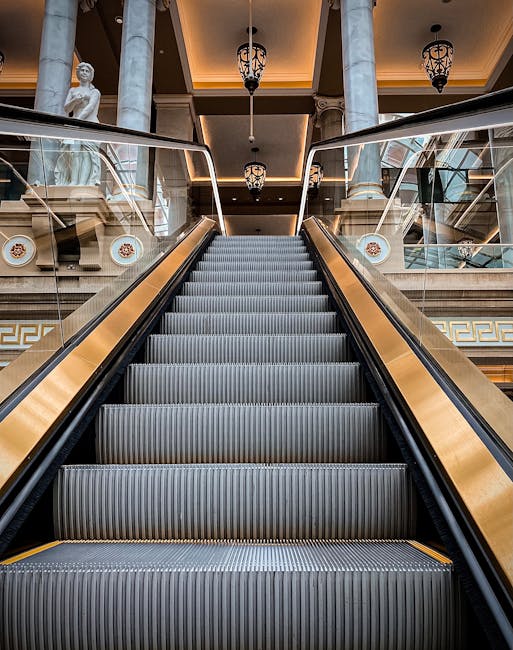 A black metal lift with a wire mesh design is positioned at the base of a staircase inside a property, serving as an access aid for home relocation or furniture transport. The lift is partially extended with its gate open, revealing a space for goods or boxes to be moved between floors. To the left of the lift, a stone staircase with polished, light-colored steps curves upward, illuminated by natural light coming through a frosted window. The interior walls are painted in a neutral tone, and a wooden handrail runs along the staircase. The environment suggests a residential setting where professional movers, such as Knightsbridge Movers, might employ this lift and stair access for efficient packing and loading during a house removal process.