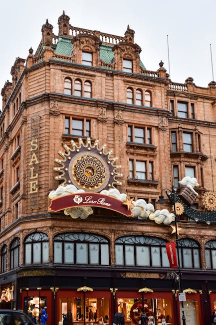 A multi-storey building in the Harrods area of Knightsbridge featuring ornate architectural details and large windows. The ground floor displays retail shop windows with mannequins and merchandise, while the upper floors have decorative stonework and tall, narrow windows. A prominent gold and white clock with sunburst rays is mounted on the facade, accompanied by a red and white banner and clouds made of foam or fabric decorations. The scene is captured during daylight with an overcast sky. This image illustrates a typical location where house removal professionals, such as Knightsbridge Movers, might conduct furniture transport and packing during a home relocation in central London, highlighting the urban environment and building's historical style.