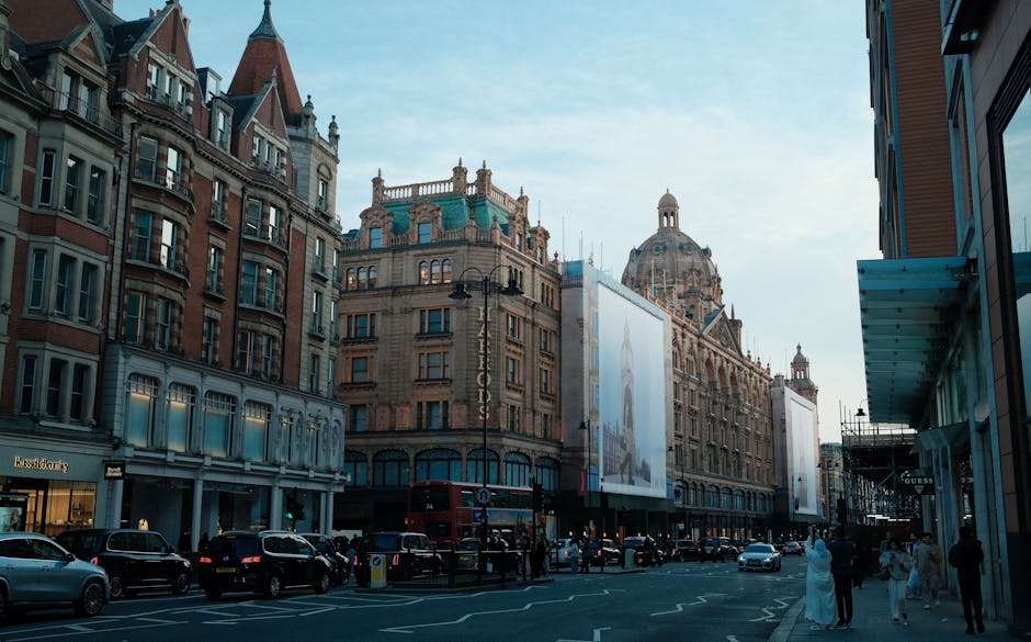 A street scene in the Harrods area of Knightsbridge, showing historic multi-storey buildings with ornate architectural details, large windows, and a domed building in the background under a clear blue sky. The street is busy with parked and moving cars along the curb, a double-decker bus in the distance, and pedestrians walking on the pavement. On the right side, a storefront with modern glass and metal elements, with a visible 'Guess' signage, indicates retail activity. The scene also features a truck or van loaded with cardboard boxes, plastic wrap, and furniture, positioned near the building entrance, with some packing materials and wrapped items visible on the pavement, suggesting a home relocation or furniture transport process managed by Knightsbridge Movers as part of a comprehensive removals service in the area.