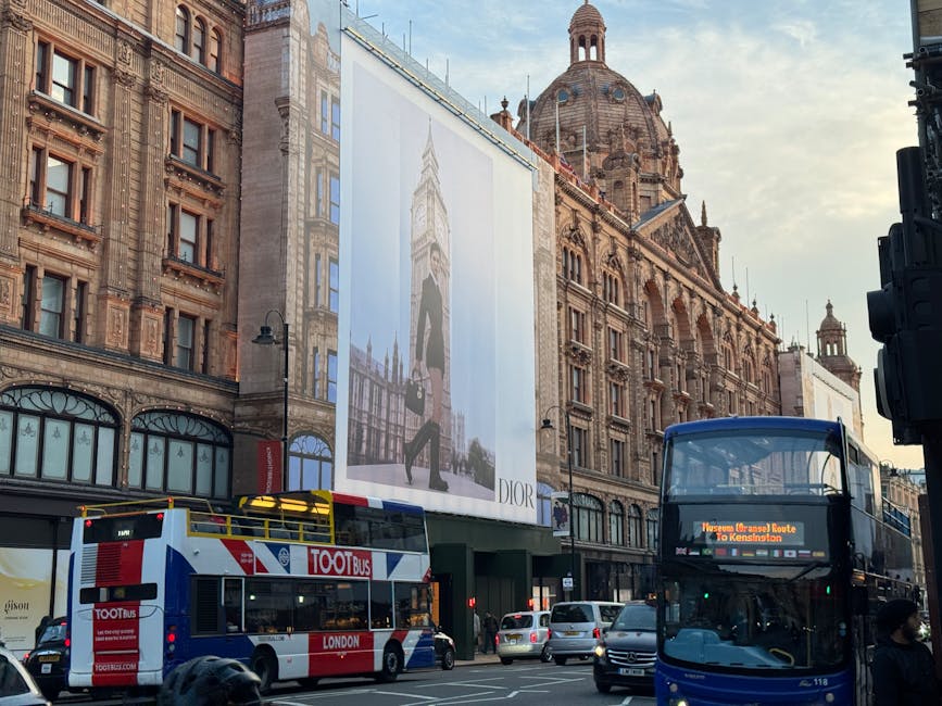 A multi-storey building in the Harrods area of Knightsbridge featuring ornate architectural details and large windows. The ground floor displays retail shop windows with mannequins and merchandise, while the upper floors have decorative stonework and tall, narrow windows. A prominent gold and white clock with sunburst rays is mounted on the facade, accompanied by a red and white banner and clouds made of foam or fabric decorations. The scene is captured during daylight with an overcast sky. This image illustrates a typical location where house removal professionals, such as Knightsbridge Movers, might conduct furniture transport and packing during a home relocation in central London, highlighting the urban environment and building's historical style.